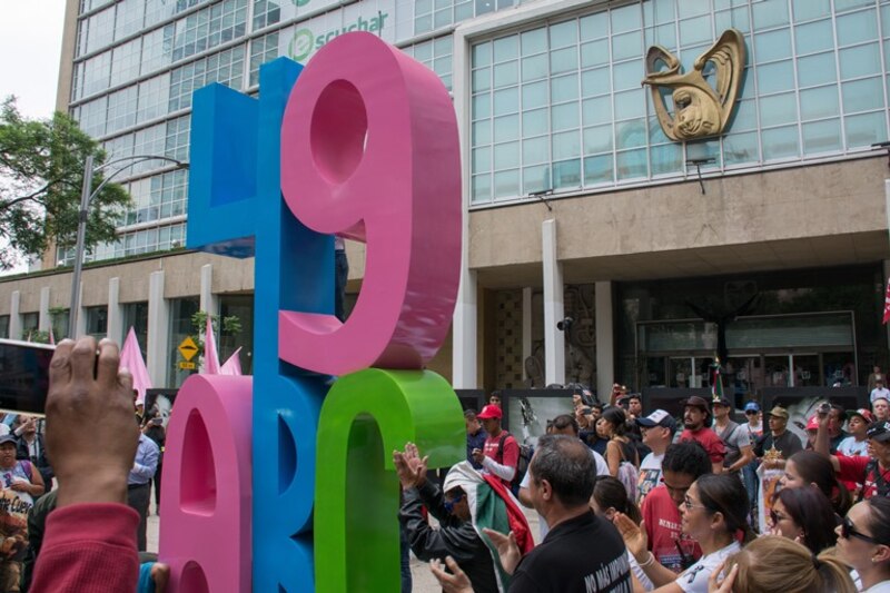 Multitud de personas en la calle mirando una escultura con forma de letras mayúsculas y de colores vivos, con un edificio de fondo y un cielo azul con nubes blancas