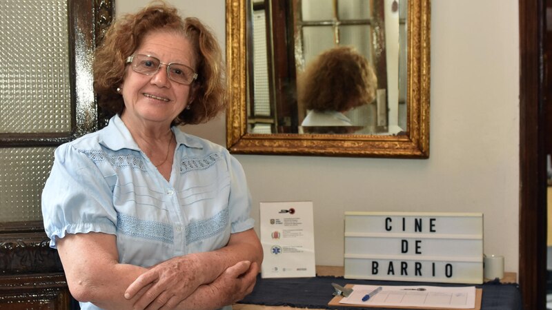 Una mujer mayor sonríe frente a un cartel que dice "Cine de Barrio".