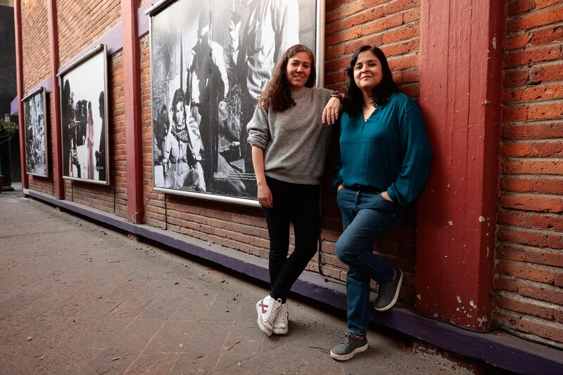 Dos mujeres sonríen apoyadas en una pared de ladrillos.