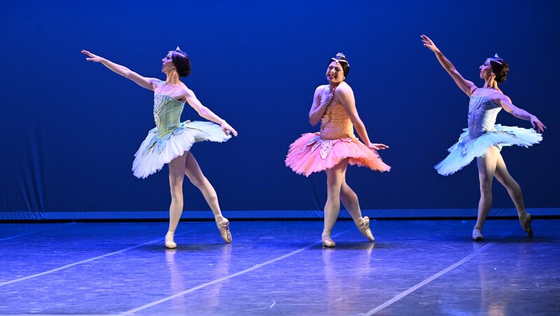 Tres bailarinas de ballet en tutús realizan una coreografía sobre un escenario con fondo azul.