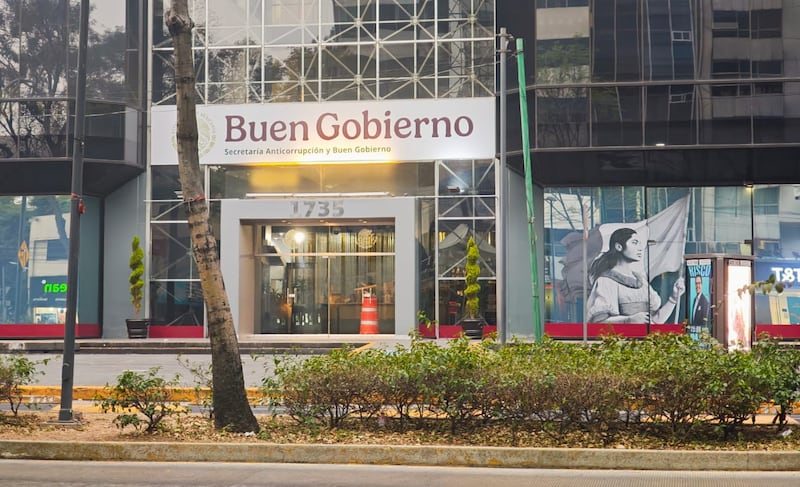 Fotografía de la entrada principal del edificio de la Secretaría Anticorrupción y Buen Gobierno en la Ciudad de México. La imagen muestra el letrero oficial con el logotipo del Gobierno de México y el nombre de la institución sobre una estructura de cristal moderna. En el ventanal derecho se aprecia un mural artístico de una mujer sosteniendo la bandera mexicana. La toma incluye elementos urbanos como árboles, una jardinera frontal y la numeración 1735 sobre el acceso. Es una imagen representativa de las instituciones gubernamentales federales y la administración pública en la capital.