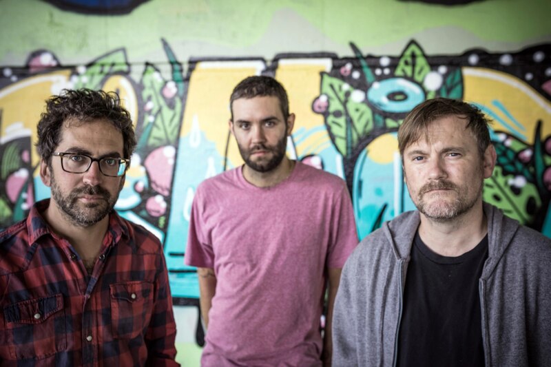 Tres hombres sonríen frente a un mural