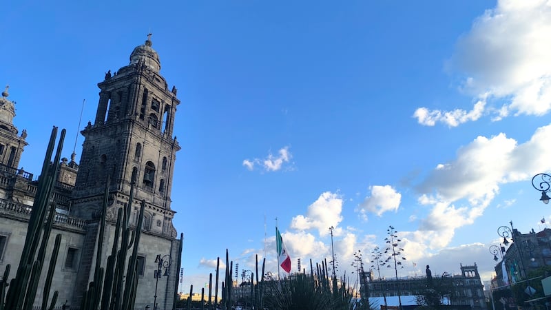 La imagen muestra una toma en ángulo contrapicado de la torre campanario de un gran edificio de piedra de estilo colonial con la bandera de México en el fondo