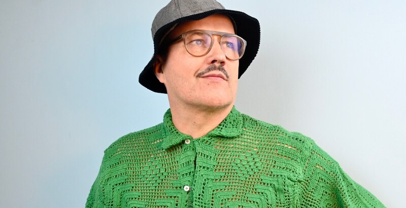 Un hombre con un sombrero de pescador verde y gafas de pasta gruesa se mira en el espejo