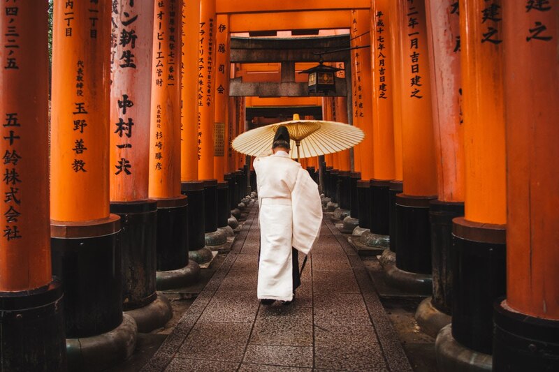 Mujer con kimono blanco y sombrilla camina bajo los arcos torii del Santuario Fushimi Inari-taisha en Kioto, Japón.