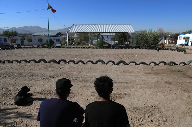 Dos hombres sentados en un campo de tierra con una hilera de llantas viejas que los separa de un perro acostado.