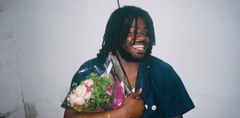 Retrato de un hombre negro con dreadlocks, barba y una sonrisa, sosteniendo un ramo de flores.