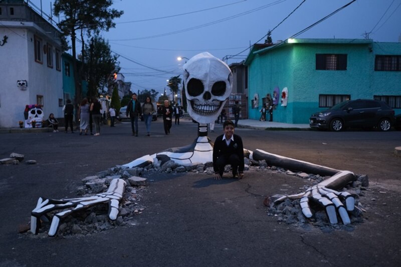 Un niño se sienta junto a una figura de un esqueleto gigante en una calle decorada con motivo del Día de Muertos.