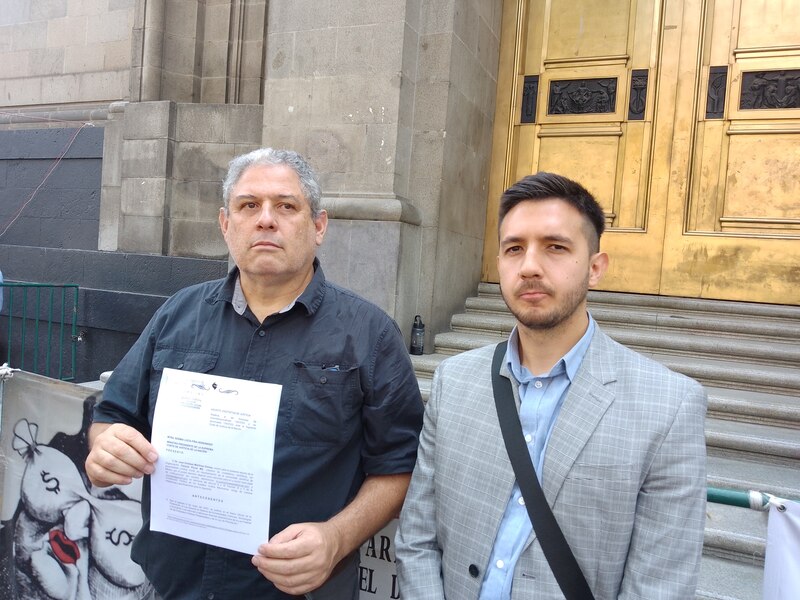 Dos hombres sostienen un cartel y un documento frente a un edificio gubernamental.