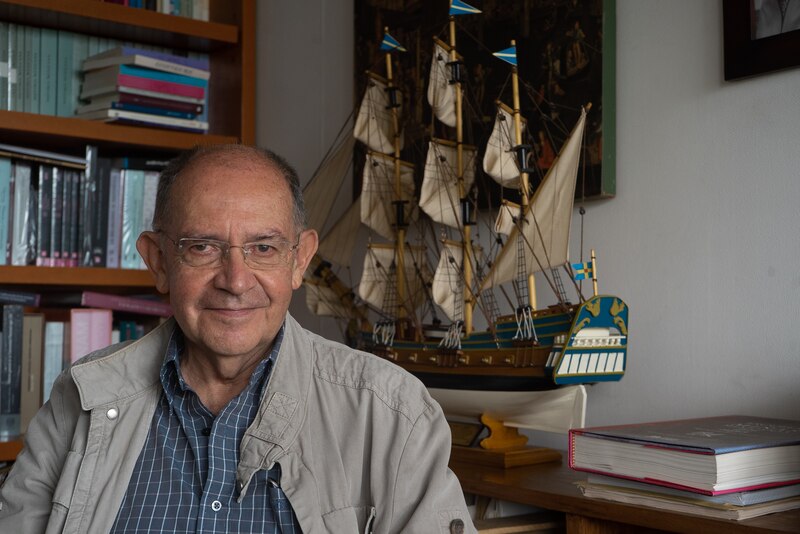 Retrato de un hombre mayor con gafas y una chaqueta marrón sentado en una silla frente a una estantería con libros y un barco modelo.