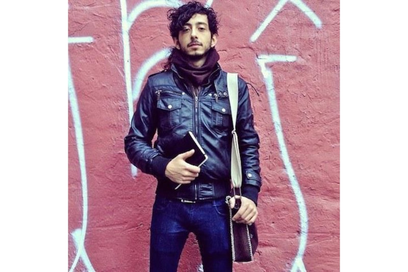 Un hombre joven con una chaqueta de cuero negra y una bufanda se encuentra de pie frente a una pared pintada con graffiti.