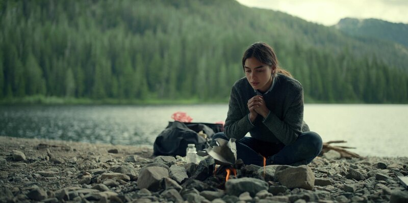 Mujer joven sentada junto a una fogata en la orilla de un lago.