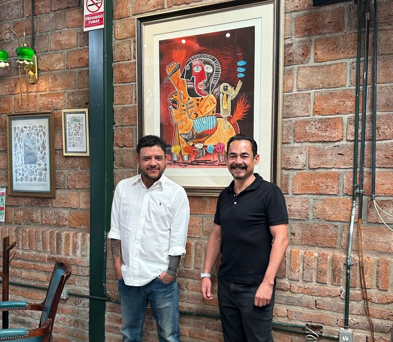Dos hombres sonríen frente a una pintura en una pared de ladrillos.