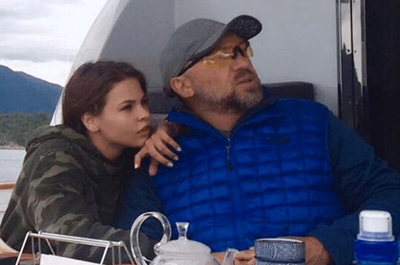 La imagen muestra a un hombre y una mujer sentados en un barco. El hombre lleva una gorra gris y una chaqueta azul, y la mujer lleva una chaqueta verde estampada. Están mirando hacia el horizonte.