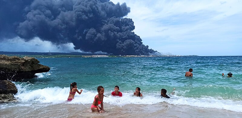 Niños jugando en la playa mientras una refinería de petróleo arde al fondo