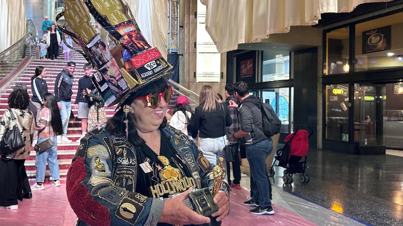 Una mujer con un sombrero extravagante y gafas de sol sostiene un Oscar en la alfombra roja