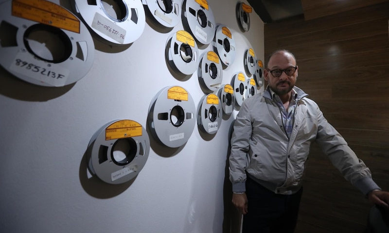 Un hombre con gafas y chaqueta gris se encuentra frente a una pared con cintas magnéticas.