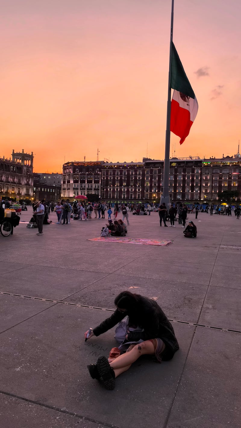 Fotografía vertical del Zócalo de la Ciudad de México durante un espectacular atardecer naranja. En primer plano, una persona sentada sobre la plancha de concreto. La Bandera Mexicana Monumental ondea en la parte superior derecha. Al fondo se distinguen los edificios del Centro Histórico de la capital, incluyendo la silueta de los portales y el Palacio Nacional. La plaza está concurrida con transeúntes. Esta imagen captura la atmósfera vibrante del Zócalo al caer la noche, combinando elementos icónicos nacionales y urbanos de CDMX.
