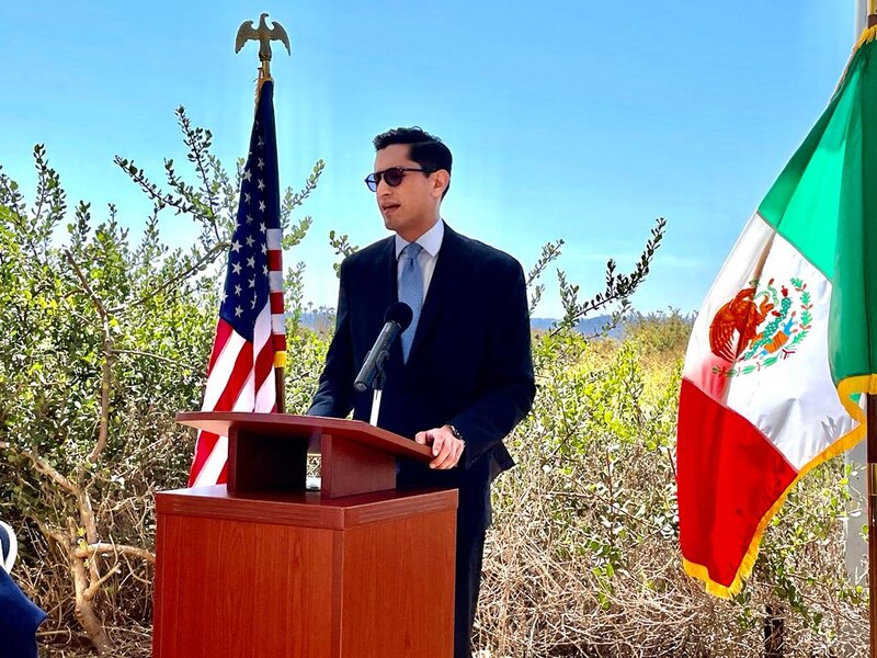El político da un discurso frente a las banderas de México y Estados Unidos.