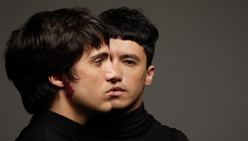 Dos hombres jóvenes con el pelo negro y las mejillas sonrosadas se miran fijamente.