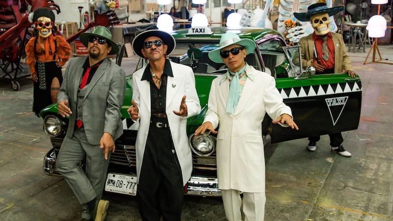 Tres hombres vestidos de blanco con sombreros y gafas de sol posan junto a un taxi verde.