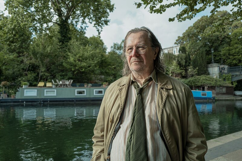 Un hombre mayor con gafas y una gabardina marrón se encuentra en un puente junto a un canal en Londres.