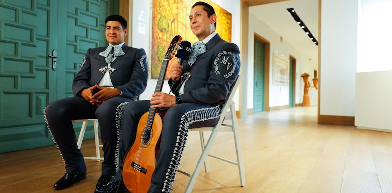 Dos hombres vestidos con trajes de mariachi están sentados en sillas y uno de ellos sostiene una guitarra.