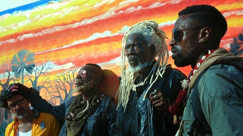 Tres hombres de raza negra con rastas y gafas de sol se encuentran de pie frente a un mural pintado en una pared.