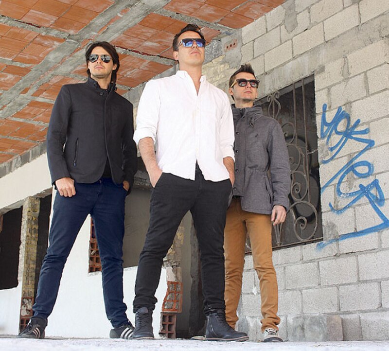 Tres hombres vestidos con ropa casual y gafas de sol posan para una foto delante de una pared de ladrillo.