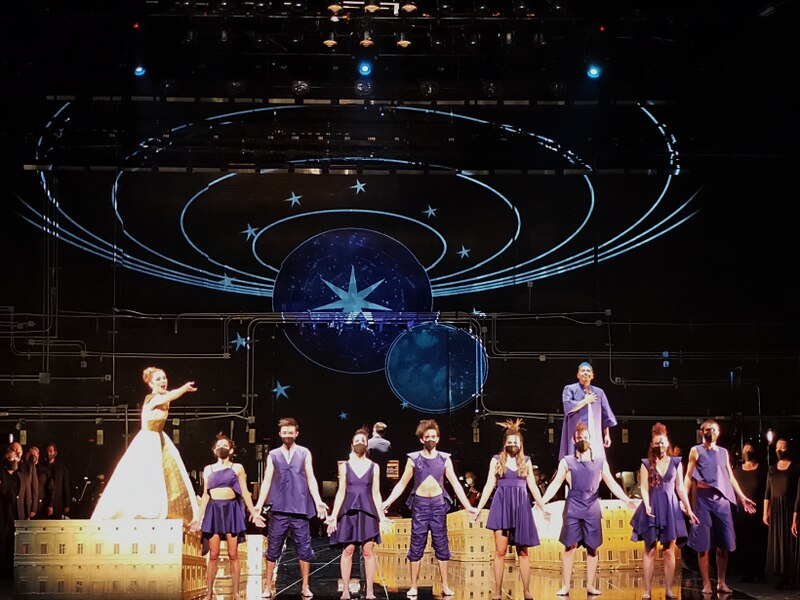 Una ópera con un coro de personas cantando y bailando en un escenario con un fondo estrellado.