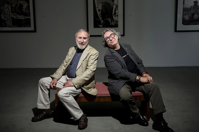 Dos hombres sonríen sentados en un banco en una galería de arte.