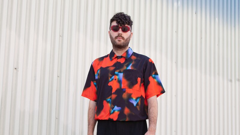 Un hombre con gafas de sol rojas y una camisa estampada se encuentra de pie frente a una pared blanca.