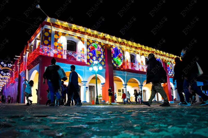 La imagen muestra a un grupo de personas caminando por una calle adoquinada en una noche estrellada. Las personas están iluminadas por las luces de Navidad que cuelgan de los edificios y los árboles.