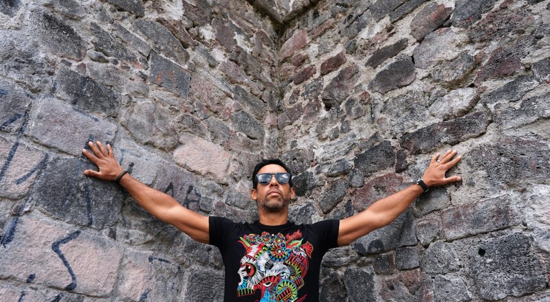 Un hombre con gafas de sol y una camiseta estampada se apoya contra una pared de piedra.