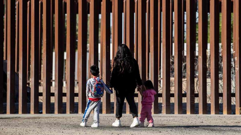 Madre e hijos migrantes caminan frente al muro fronterizo