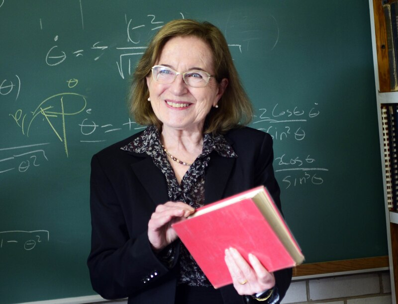 Profesora sonríe mientras sostiene un libro frente a una pizarra