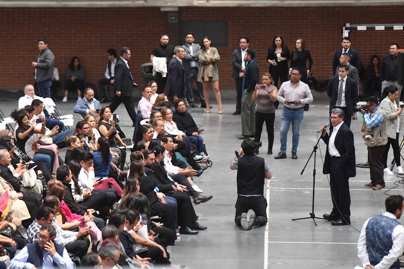 Un reportero entrevista a un político mientras un grupo de personas observa.