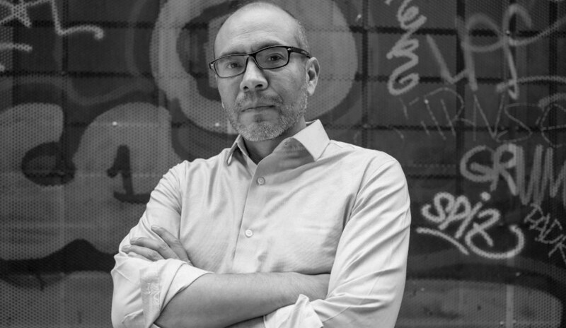 Retrato en blanco y negro de un hombre con gafas y barba delante de un muro de graffiti.