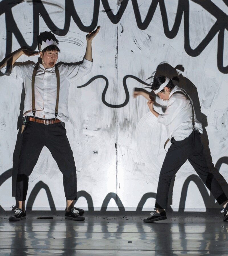 Dos bailarines realizan una coreografía contemporánea con sombras proyectadas sobre una pared blanca.