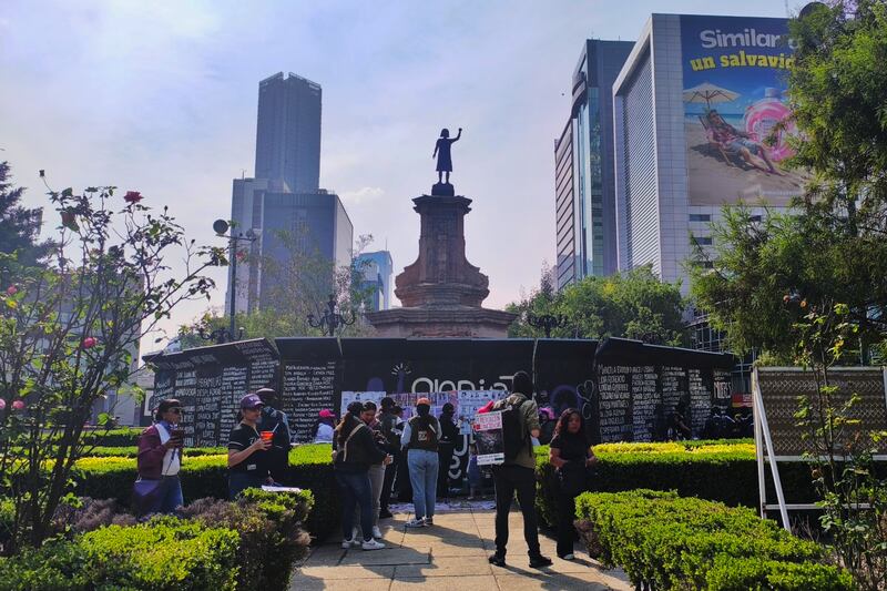 Vista diurna de la Glorieta de las Mujeres que Luchan (anteriormente conocido como Glorieta de Colón), ubicada en el Paseo de la Reforma de la Ciudad de México. La imagen captura la base del monumento con la figura central, rodeada por el cerco cubierto de mensajes y nombres de víctimas de feminicidio y desaparecidas, que sirve como un memorial feminista y espacio de protesta. Se aprecian personas observando las consignas y el entorno de jardines con arbustos bien cuidados. Al fondo, se elevan los modernos rascacielos característicos de la arquitectura del corredor financiero de Reforma. Es un punto clave que simboliza la lucha por la justicia y la memoria en la capital mexicana.