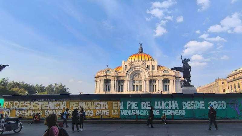 Fotografía idel Palacio de Bellas Artes en el Centro Histórico de la Ciudad de México, con su característica cúpula dorada y naranja bajo un cielo azul. En primer plano, se observa el muro o cerco temporal que protege el edificio, cubierto con una gran manta o mural de consignas feministas. El mensaje central hace referencia a la "Resistencia Feminista en pie de lucha". La imagen captura la mezcla entre la arquitectura histórica y la manifestación social contemporánea, mostrando a transeúntes y activistas en la calle. Un punto de referencia clave que subraya la intersección entre cultura, historia y movimientos sociales en la capital mexicana.