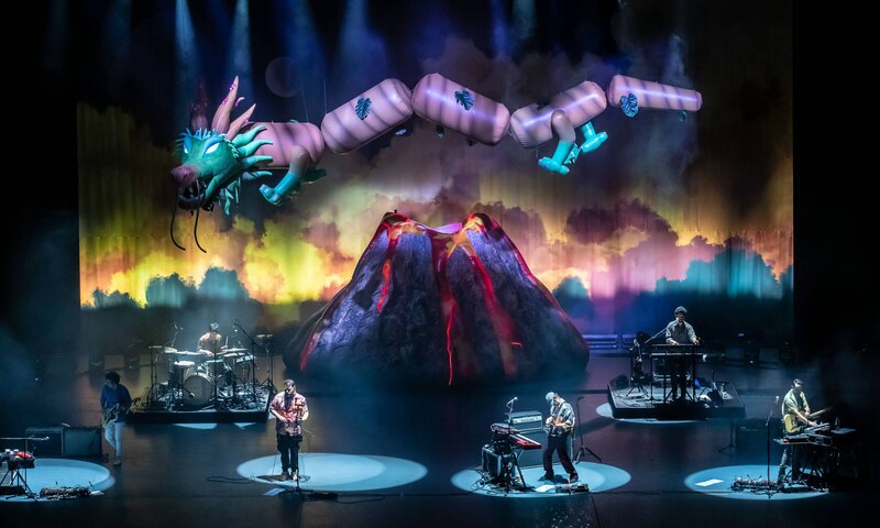 Banda de música tocando en vivo en un escenario con un dragón inflable gigante y un volcán en el fondo.